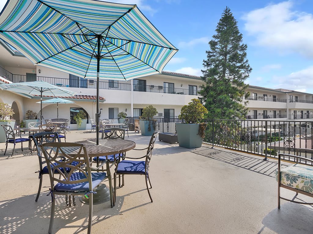 A sunny outdoor patio area with tables, chairs, and large umbrellas by a multi-story building.