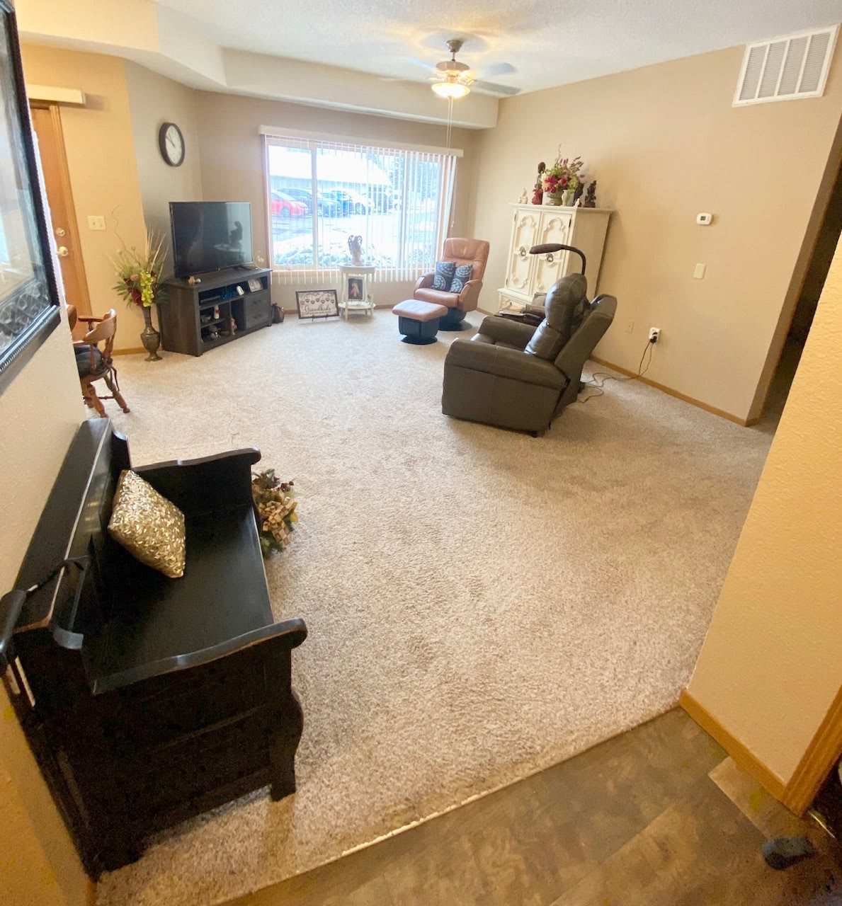 A cozy living room with comfortable seating and a television, featuring neutral tones and ample natural light from a large window.