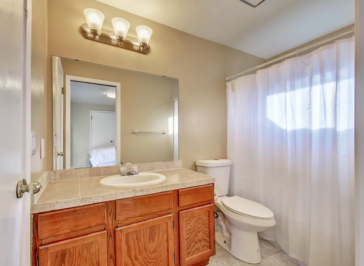 A well-lit bathroom featuring a wooden vanity, sink, and toilet with a white shower curtain.