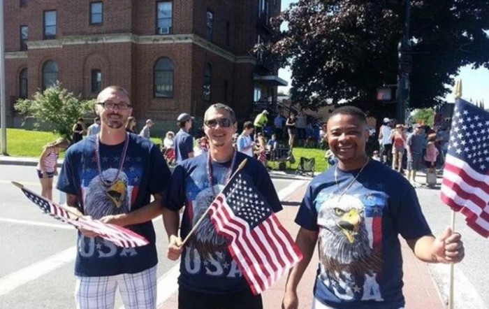 Three people wearing patriotic clothing participate in a parade, holding American flags on a sunny day.