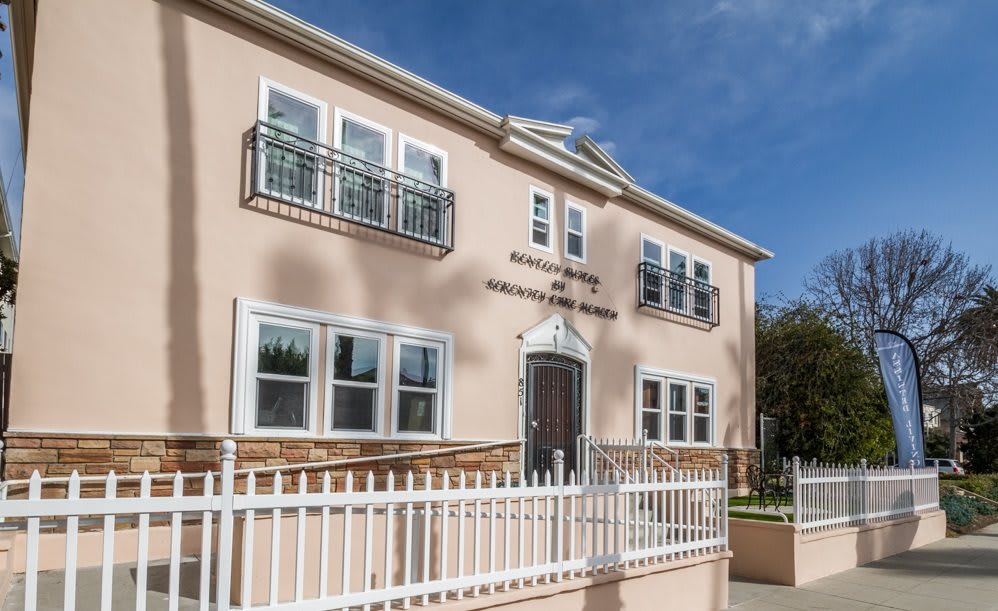 A two-story house with a light pink facade, featuring multiple windows and a white fence in the foreground.