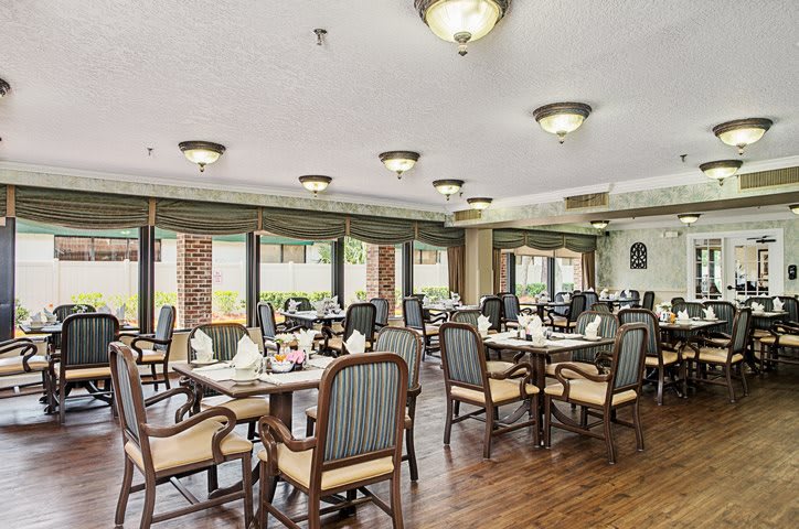 A well-lit dining area with elegantly arranged tables and chairs, featuring large windows and wooden flooring.