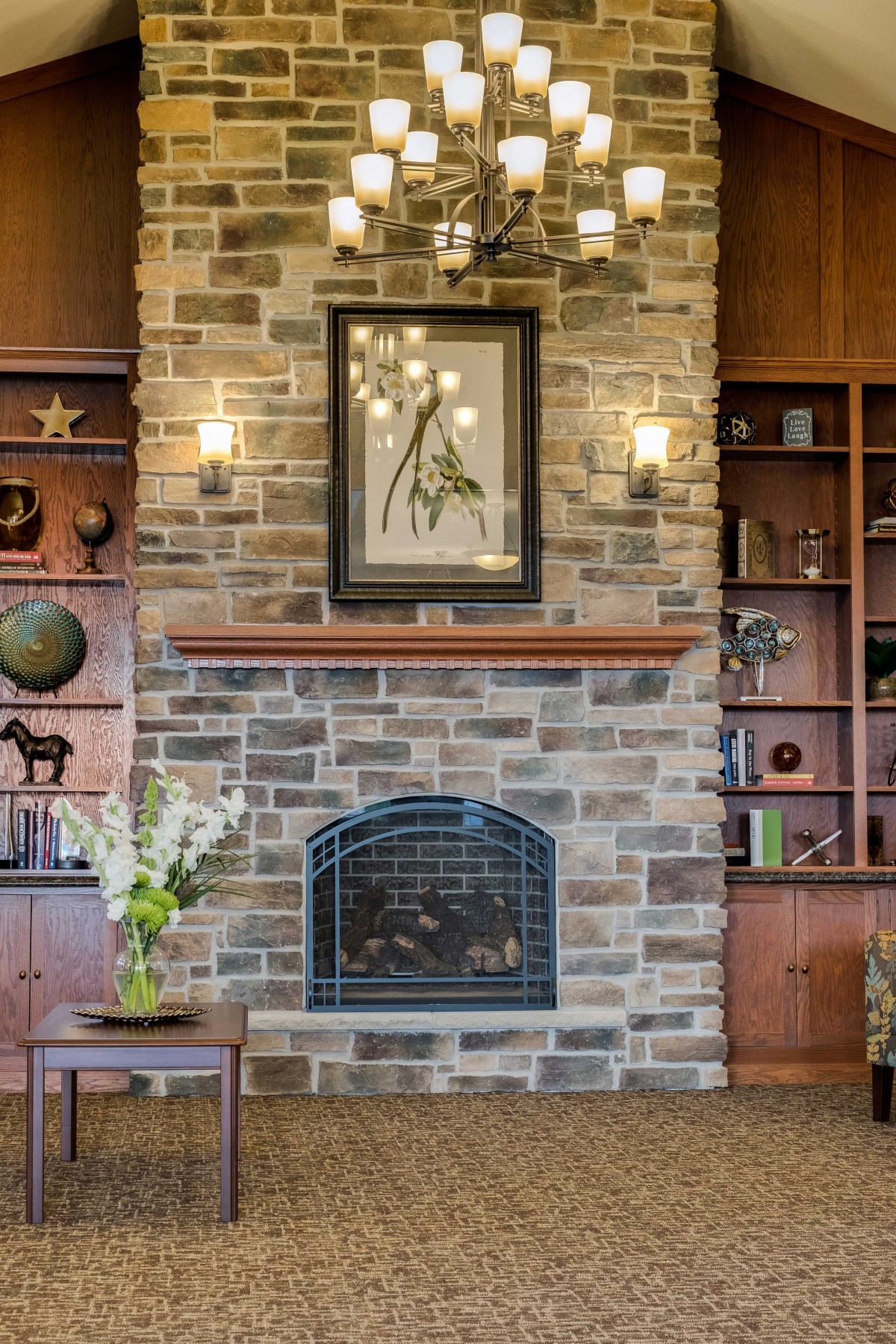 A cozy living room features a stone fireplace, flanked by wooden bookshelves adorned with various decorative items and artwork. A vase of flowers adds a touch of nature to the setting.