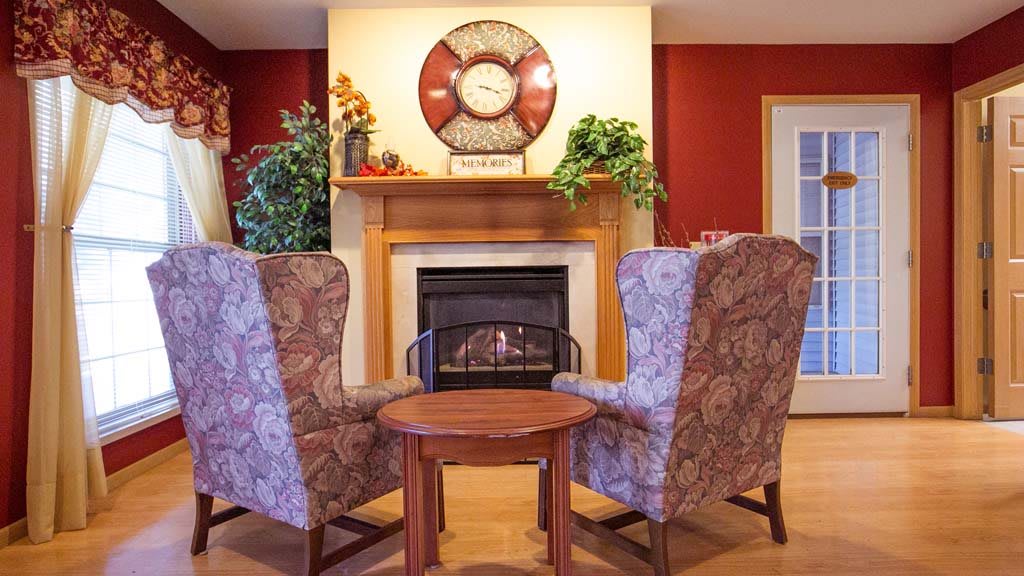 A cozy living room featuring a warm fireplace, two patterned chairs, and a small round table.