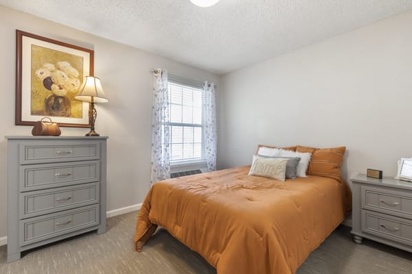 A cozy bedroom with an orange bedspread, a dresser with a lamp, and a window with sheer curtains.