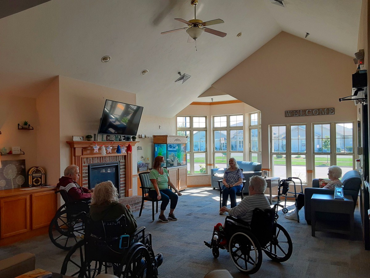 A group of seniors sitting in a communal living room, some in wheelchairs, engaged in a social gathering.
