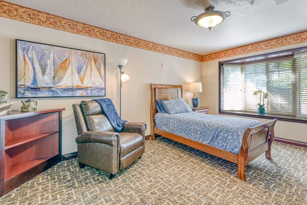 A cozy bedroom featuring a bed, armchair, and nautical-themed artwork, with ample natural light from a large window.