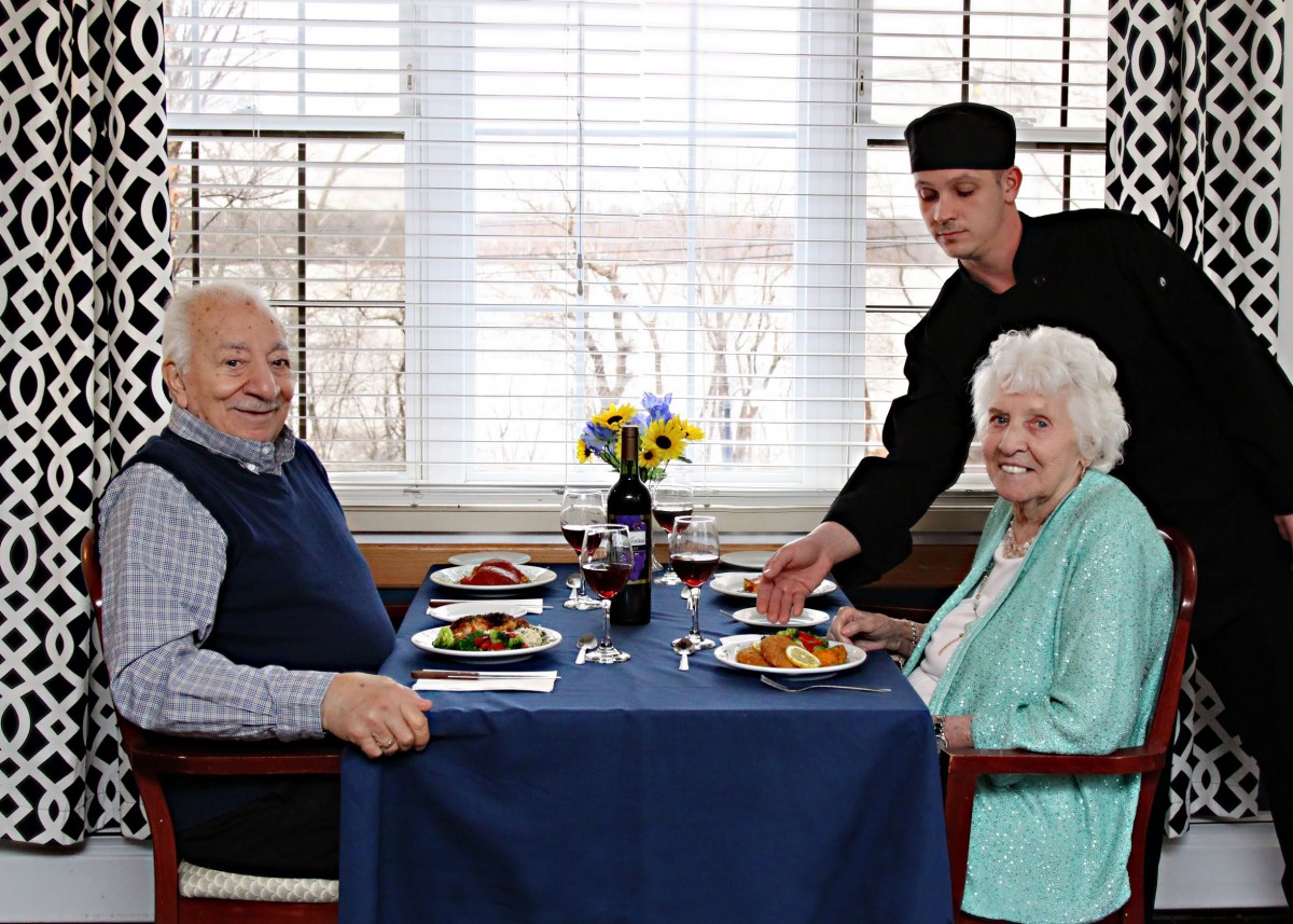 An elderly couple enjoying a meal at a restaurant, served by a waiter.