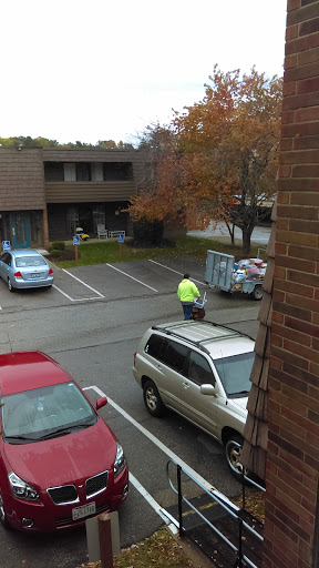 A worker in a bright yellow vest collects trash in a parking lot with autumn foliage.
