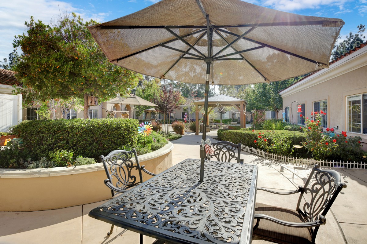 A sunny patio with a metal table and chairs under a large umbrella, surrounded by garden greenery.