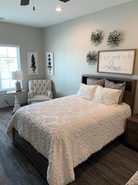 A modern and cozy bedroom with a neatly made bed, an upholstered chair, wall art, and a lampshade by the window.