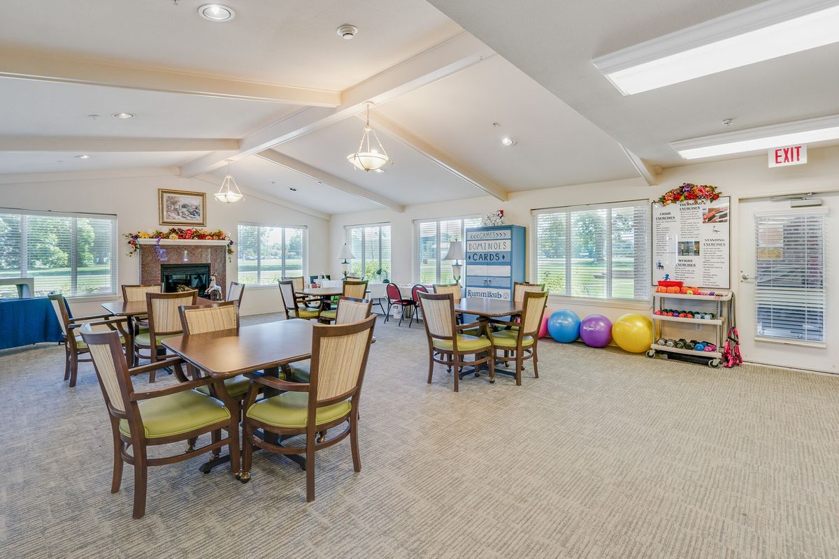 A spacious community room with tables, chairs, and windows, featuring exercise equipment in the corner.