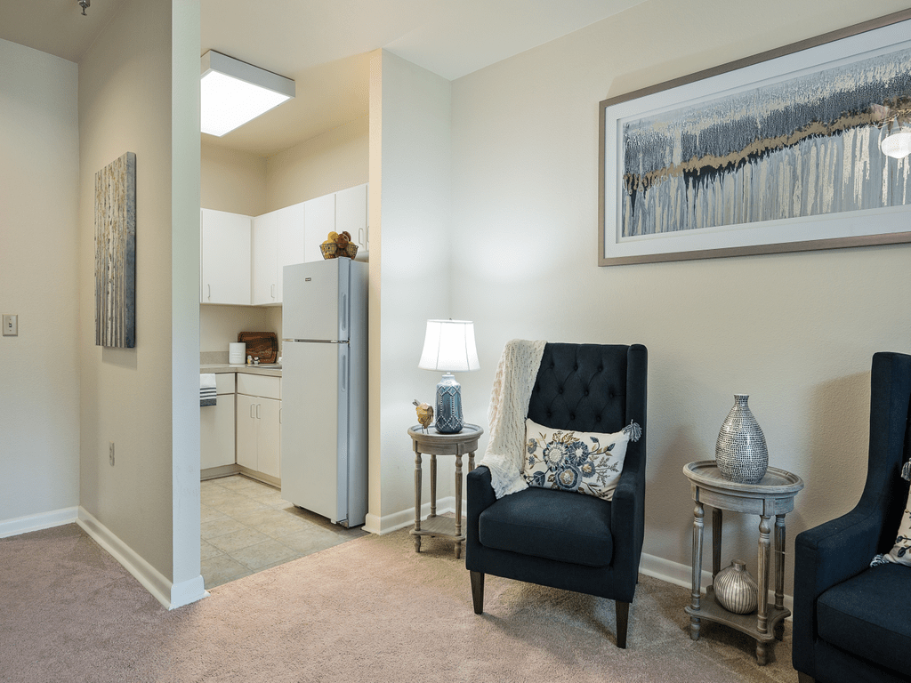 A cozy living space adjacent to a small kitchen area, featuring a comfortable black chair, side tables with decor items, and wall artwork.