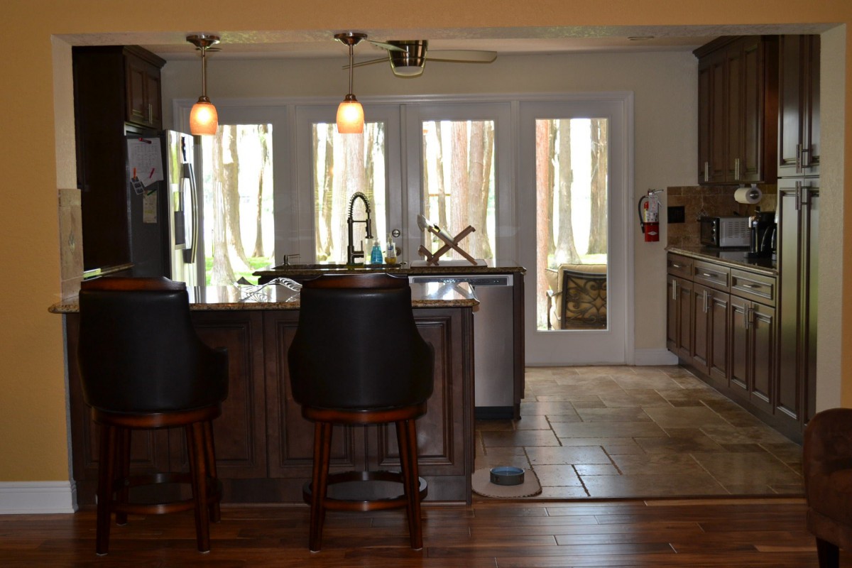 A modern kitchen interior featuring bar stools, wooden cabinets, and large windows offering natural light.