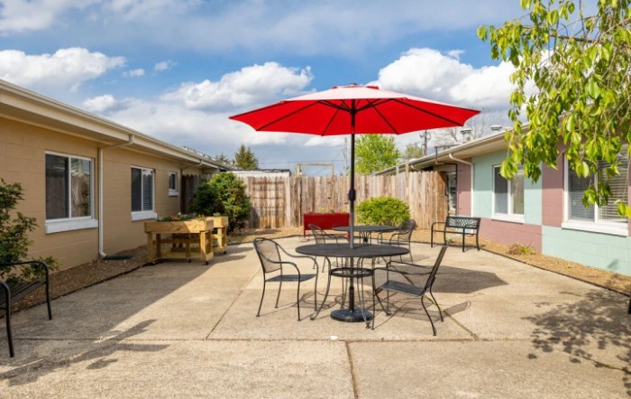 A sunny backyard patio with a red umbrella over a table and chairs.