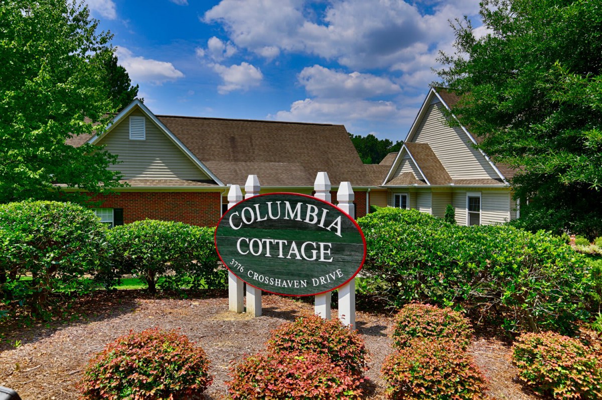 Entrance sign of Columbia Cottage surrounded by greenery under a partly cloudy sky.