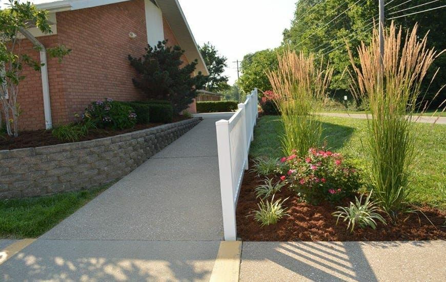 A well-maintained garden alongside a concrete pathway with a white fence next to a brick building.
