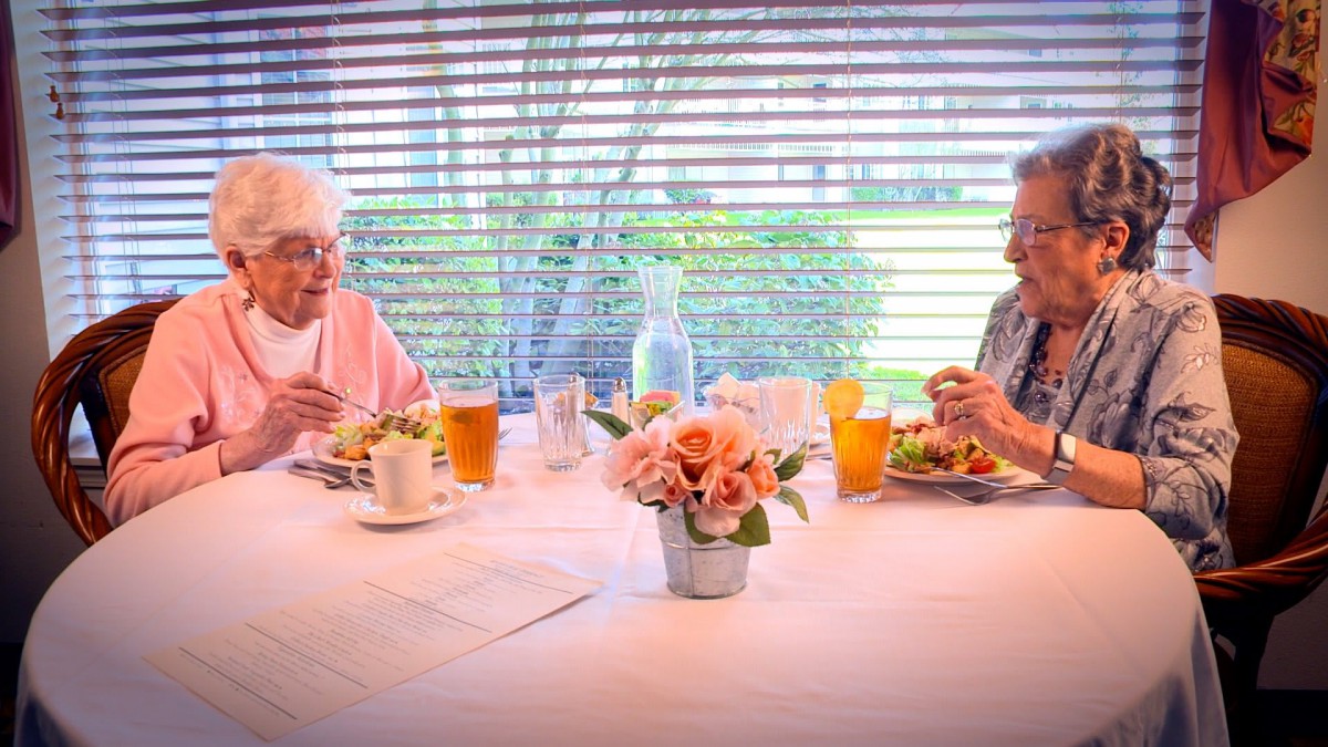Two elderly people enjoying a meal together at a table with flowers and drinks.