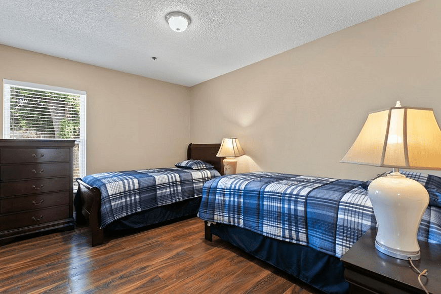 A cozy bedroom featuring two twin beds with blue plaid bedding, a lamp on a nightstand, and wooden flooring.