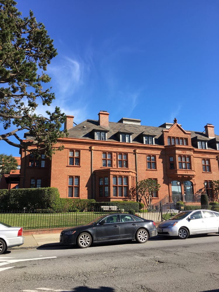 A historic brick mansion with multiple chimneys and dormer windows, set against a clear blue sky, with parked cars in front.