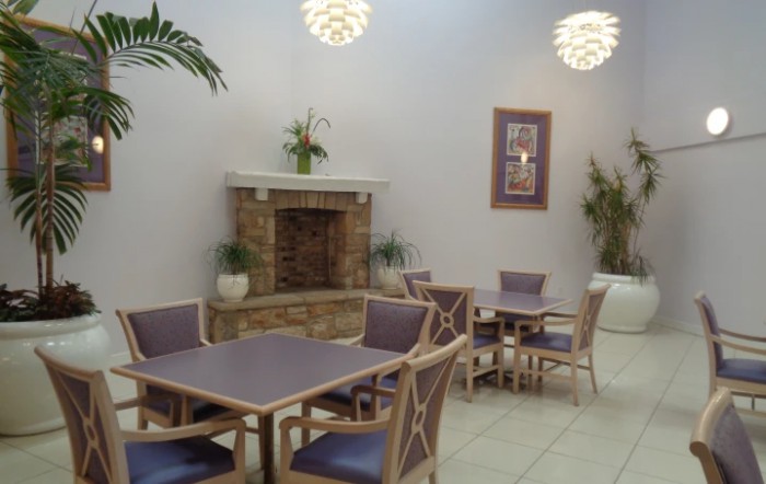 A cozy indoor seating area with tables and chairs near a fireplace, adorned with plants and soft lighting.