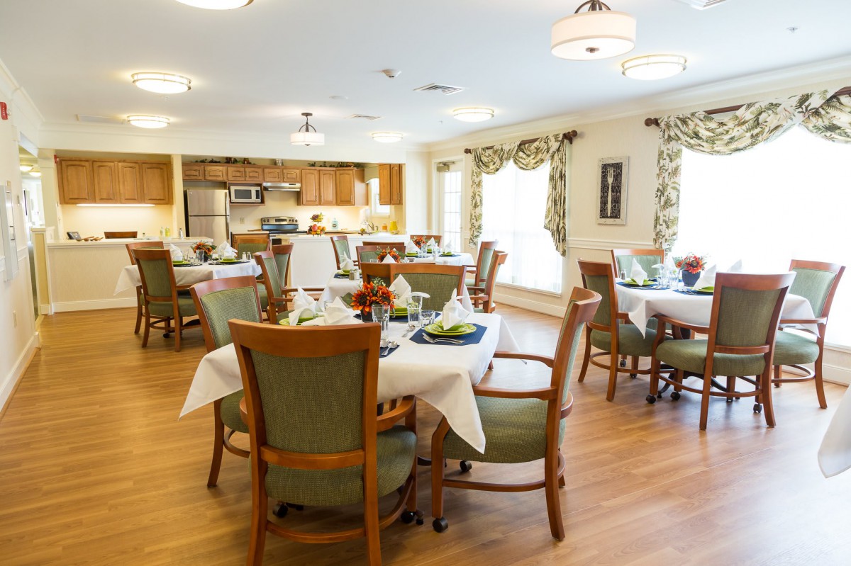 A well-lit dining room with multiple tables and chairs arranged neatly, featuring a kitchen area in the background.