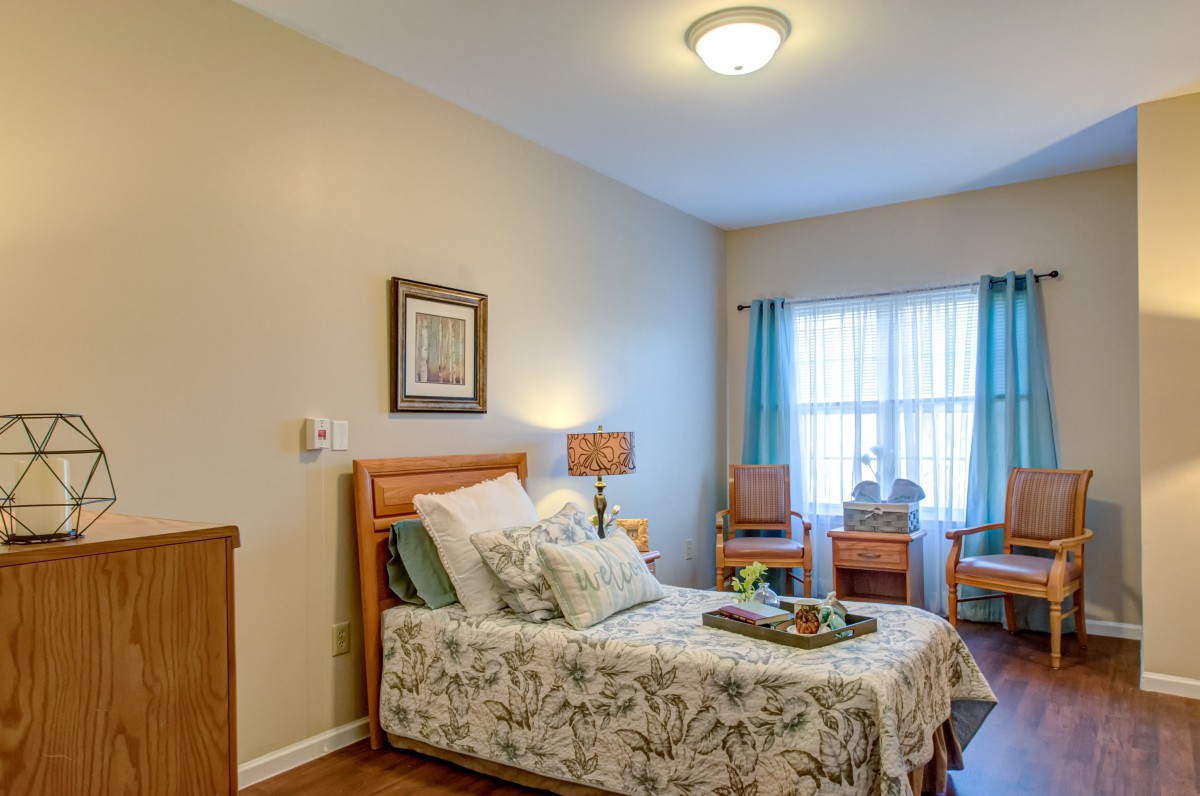 A cozy and well-lit bedroom featuring a single bed with floral bedding, a lamp, and natural light streaming through the window.