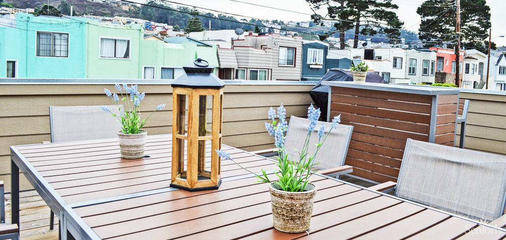 A rooftop deck with a wooden table featuring a lantern and potted plants, overlooking an urban neighborhood.