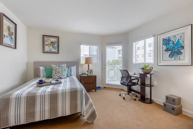A cozy bedroom with a neatly made bed, a desk with a chair, and several windows allowing natural light to brighten the space.