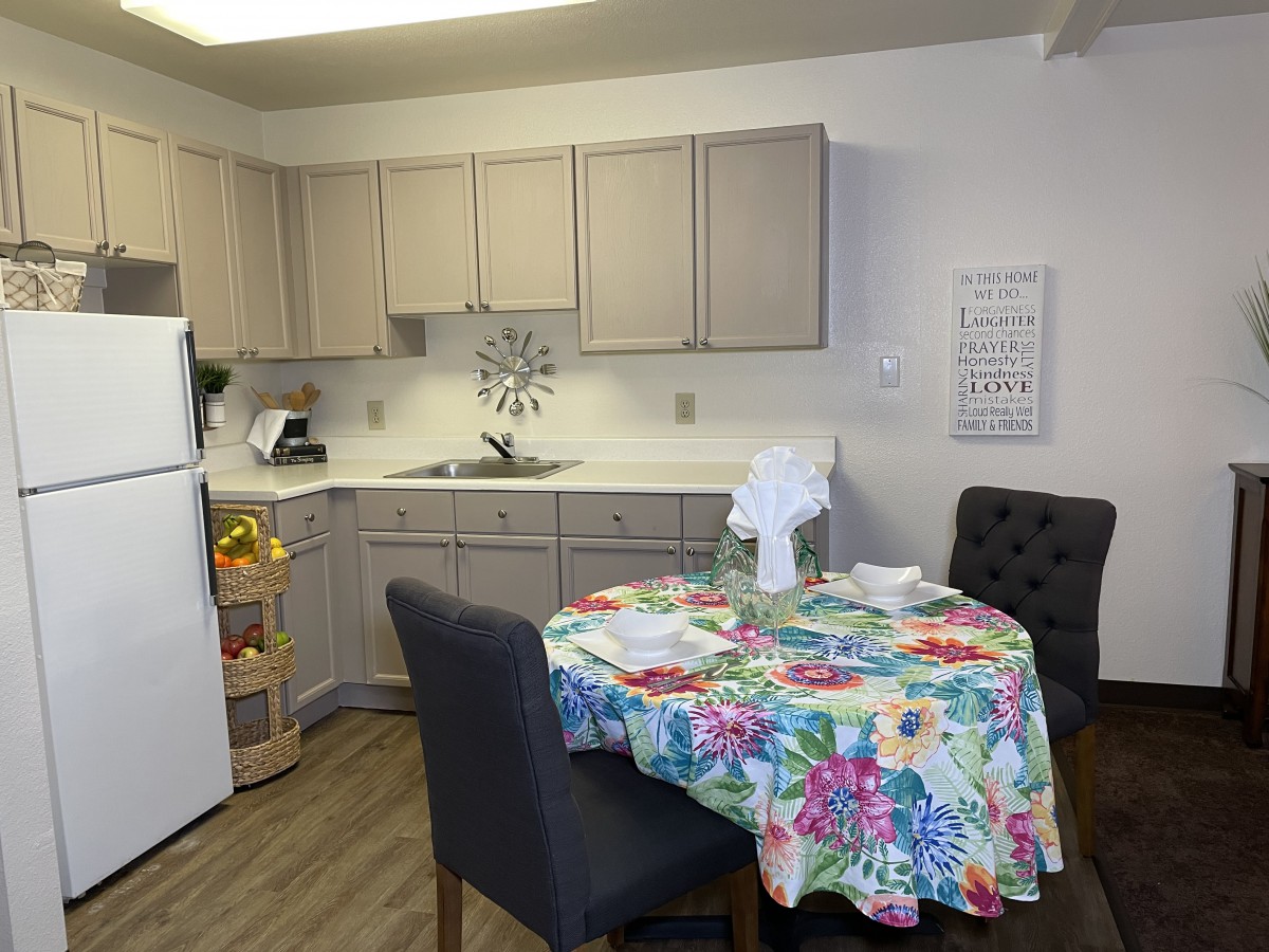 A cozy kitchen and dining area featuring a table with a colorful floral tablecloth, surrounded by chairs, with cabinets and kitchen appliances in the background.