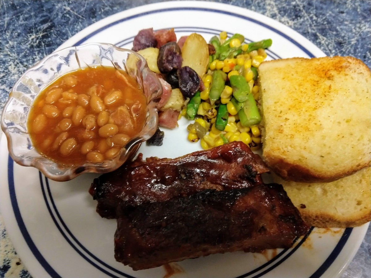 A dinner plate featuring ribs, beans, roasted potatoes, corn and asparagus mix, and a slice of bread.