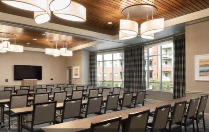 A modern conference room with rows of tables and chairs, featuring large windows and stylish lighting.