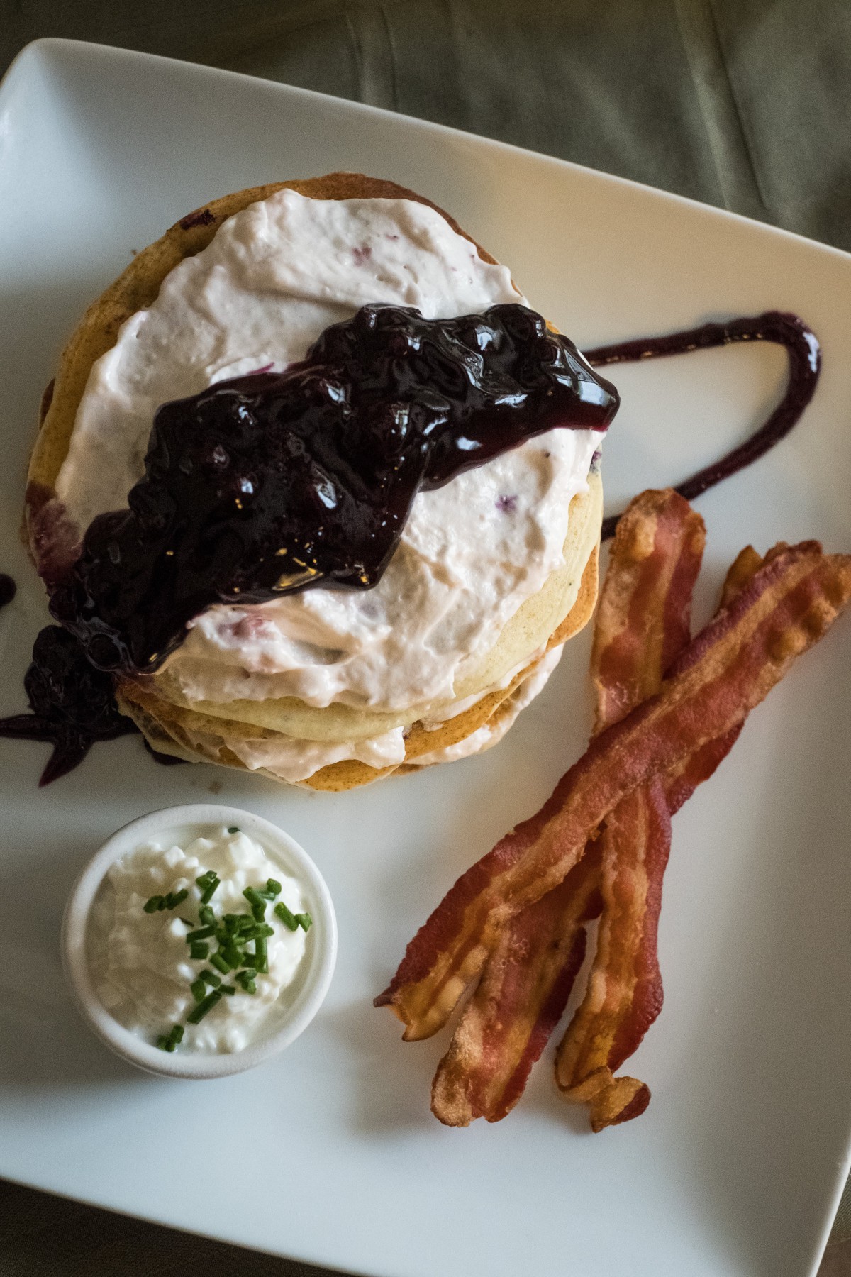 A delicious breakfast featuring pancakes topped with blueberry sauce, served with crispy bacon and a side of whipped cream garnished with chives.