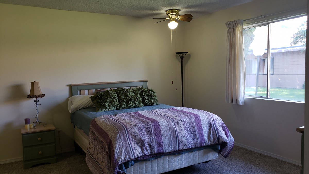 A cozy bedroom featuring a neatly made bed, ceiling fan, standing lamp, and window with curtains.
