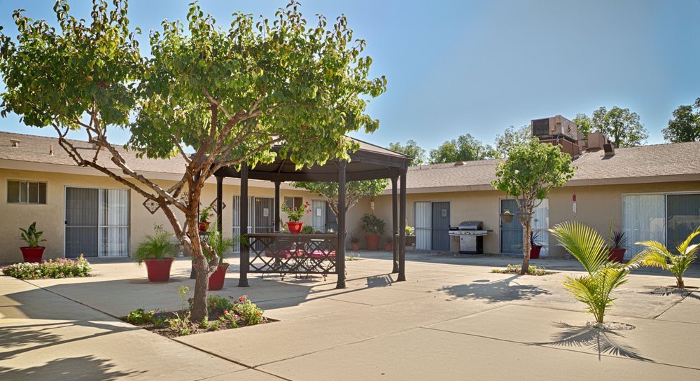 A sunny courtyard featuring a gazebo with outdoor seating, surrounded by trees and potted plants.