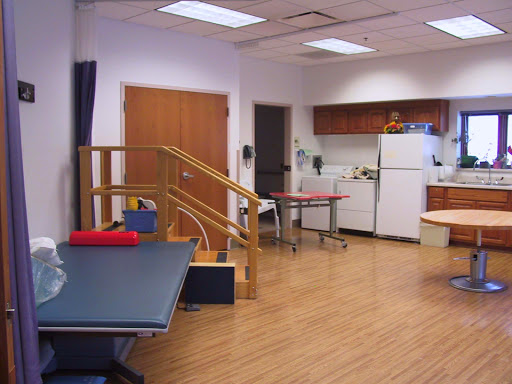 A well-equipped rehabilitation therapy room featuring exercise tables, a staircase for mobility training, and a kitchenette.
