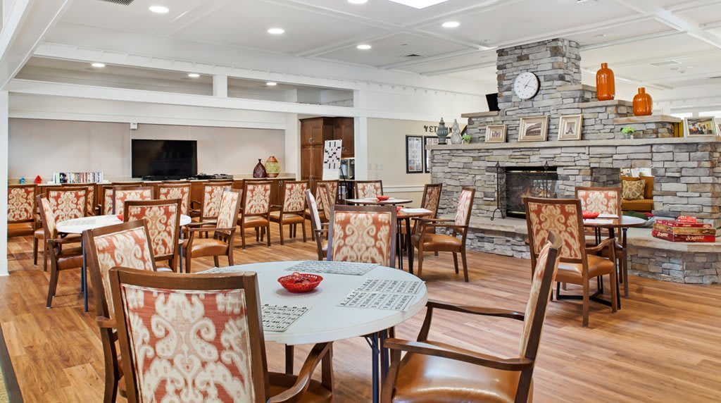 A cozy dining area with patterned chairs and round tables, featuring a stone fireplace and warm wooden flooring.