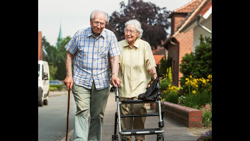 An elderly couple enjoys a walk together on a sunny day, with the woman using a walker for support.
