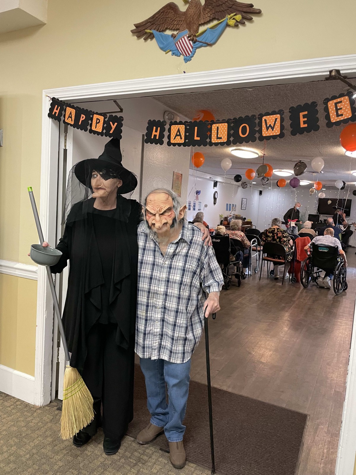 Two people in Halloween costumes stand under a 'Happy Halloween' banner at a festive indoor gathering.