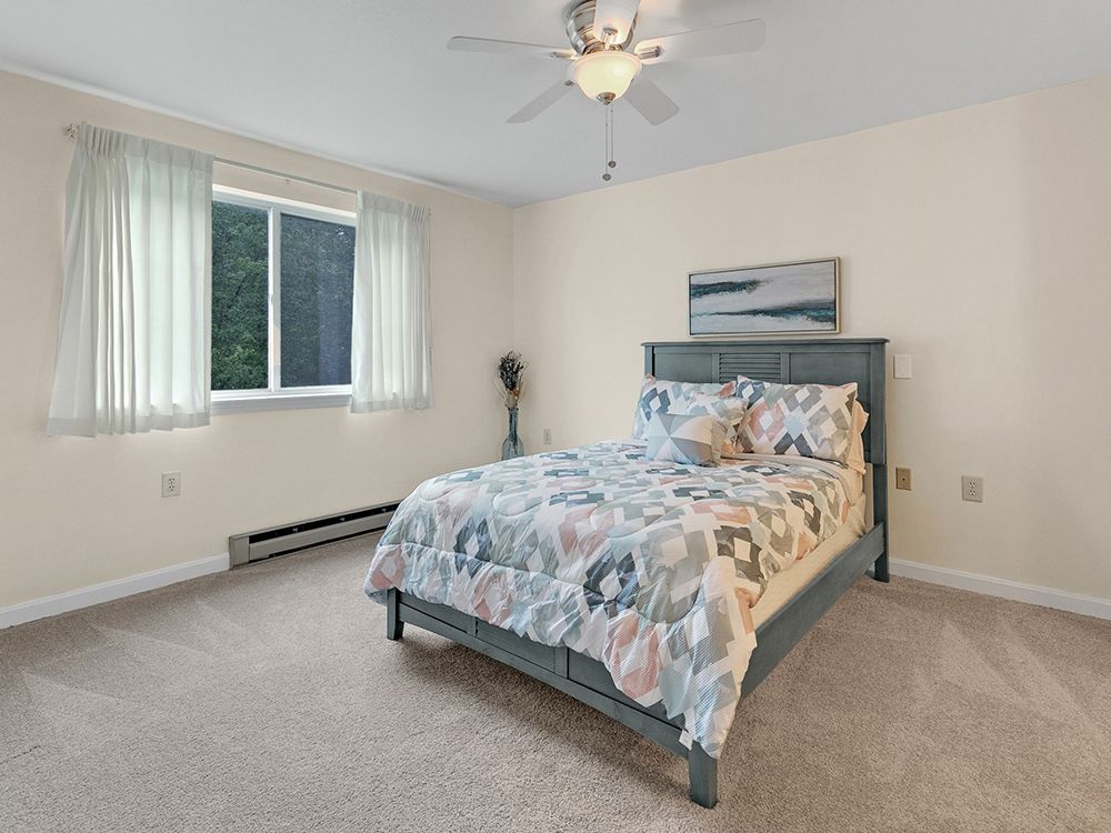 A cozy bedroom with a bed covered in a patterned duvet, a window with curtains, and a ceiling fan.