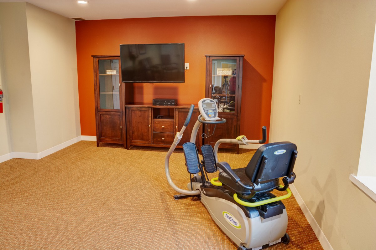 A cozy exercise room featuring a recumbent bike and a television, with wooden cabinets against an orange accent wall.