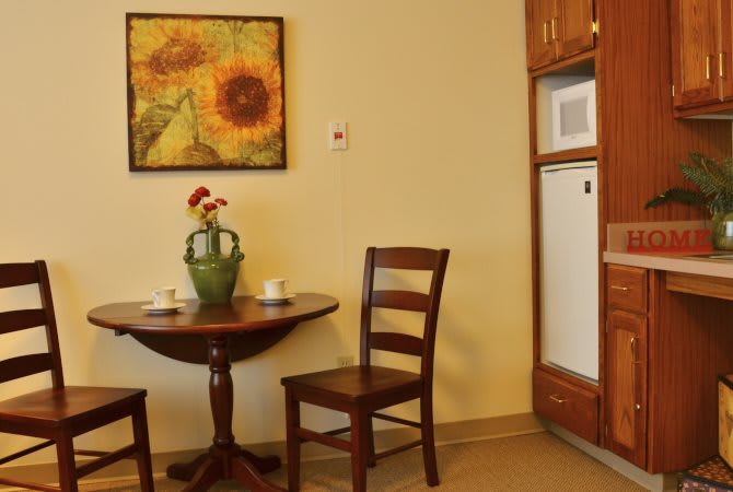 A cozy kitchen corner with a small round table, two wooden chairs, a vase with flowers, and sunflower artwork on the wall.