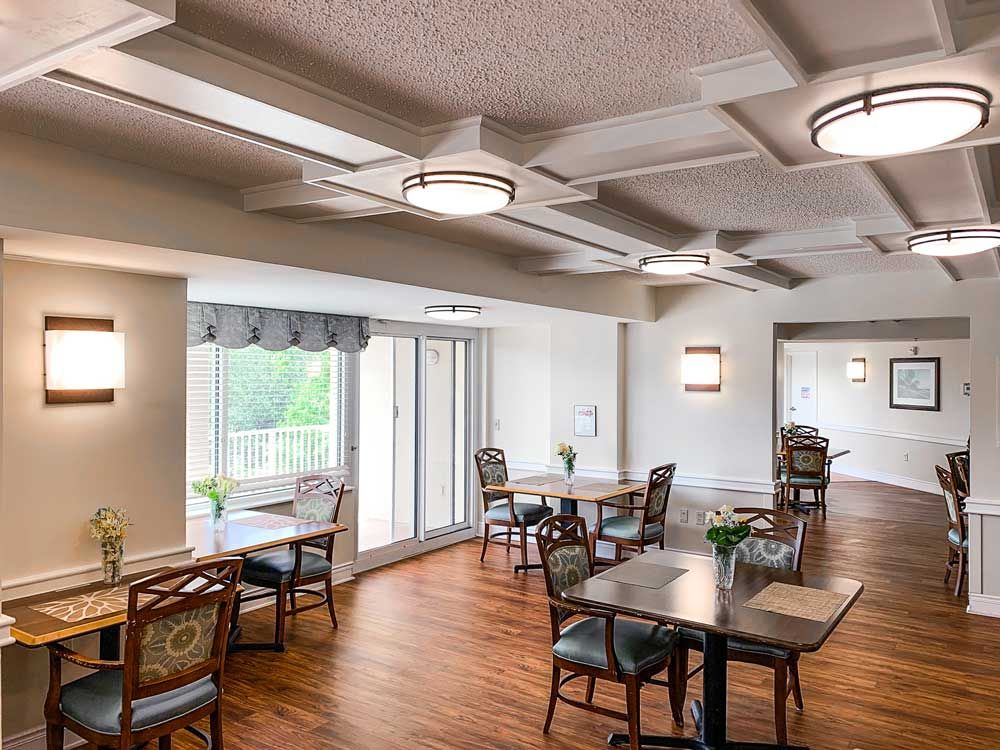 A well-lit dining room with multiple tables and chairs, featuring wood flooring and large windows.