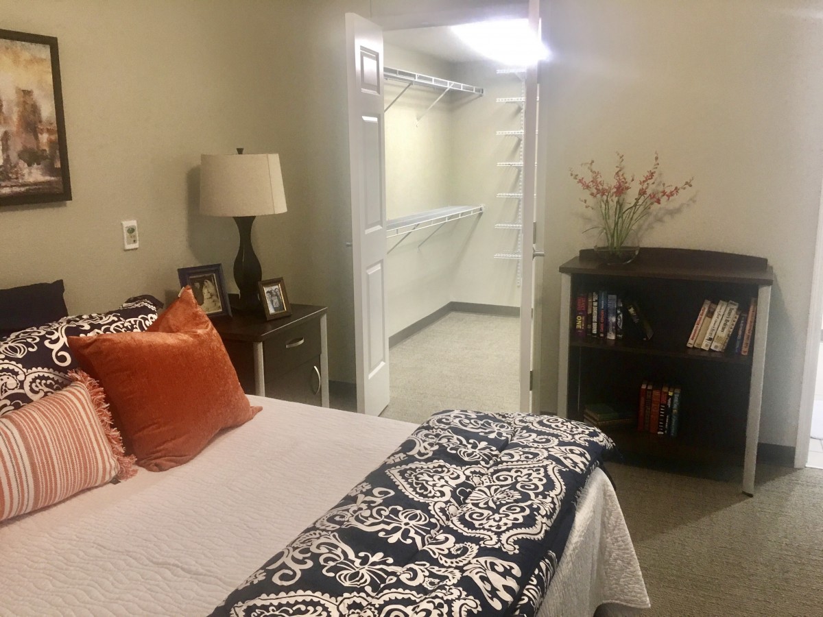 A cozy bedroom with a neatly made bed, featuring vibrant pillows and a patterned throw. A lamp sits on the bedside table, and a bookshelf is stocked with books. The room opens to a walk-in closet.