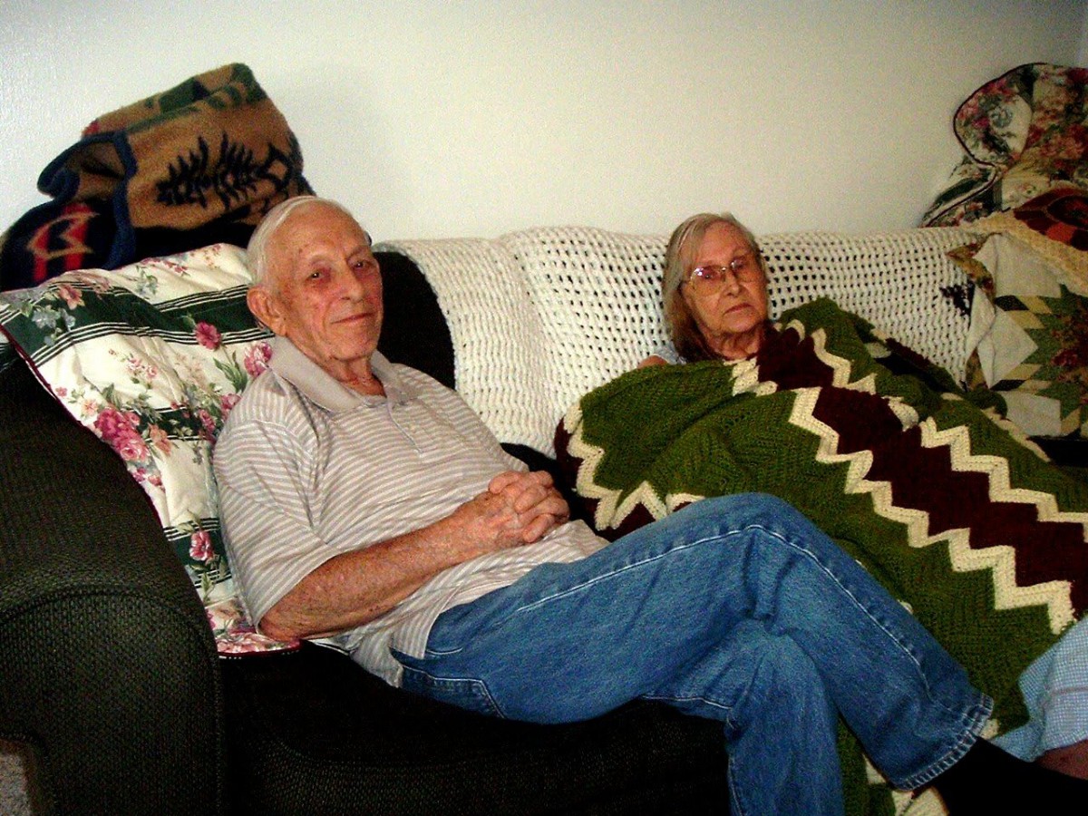 An elderly couple sits comfortably on a couch, with one of them wrapped in a blanket.