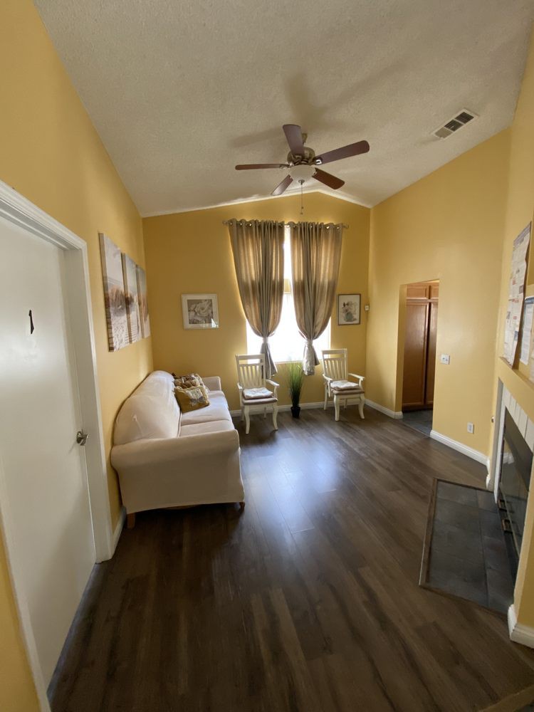A cozy living room with a sofa, curtains, and a ceiling fan, featuring warm yellow walls and wooden flooring.