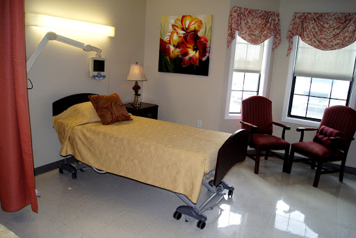 A well-lit hospital room featuring a single bed with yellow bedding, a comfortable chair, and decorative artwork on the wall.