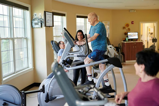 An elderly man engages in a workout on an elliptical machine with the guidance of a personal trainer at a fitness center.