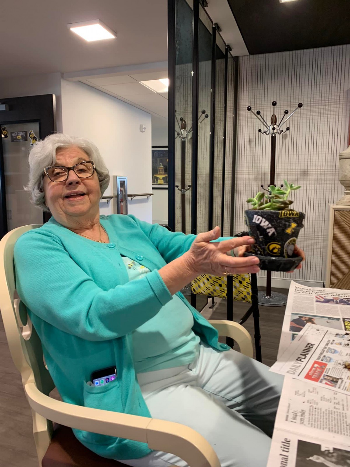 An elderly woman with glasses is smiling while sitting indoors, holding a plant.