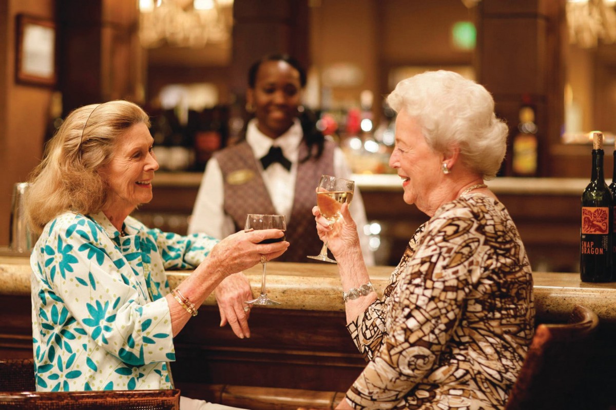 Two elderly women enjoy a toast at a bar, sharing laughter in a warm and social setting.
